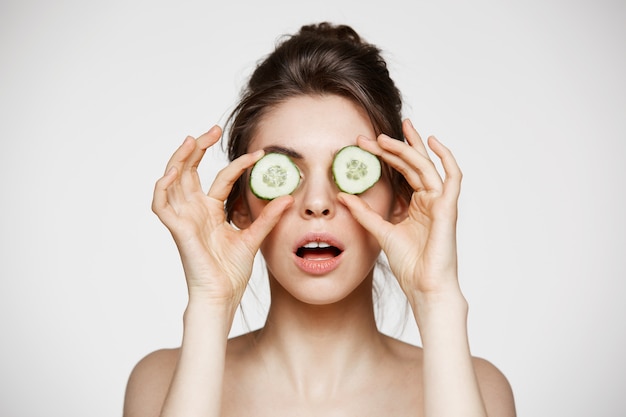 Jeune belle fille nue avec la bouche ouverte, cachant les yeux derrière des tranches de concombre sur fond blanc. Soins de beauté et cosmétologie.