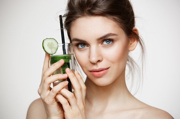 Jeune belle fille avec une peau parfaitement propre souriant regardant la caméra tenant un verre d'eau avec des tranches de concombre sur fond blanc. Alimentation saine.