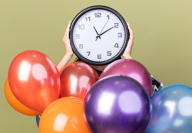 Jeune belle fille portant un chapeau bleu debout derrière des ballons recouverts d'une horloge murale isolée sur un mur vert olive