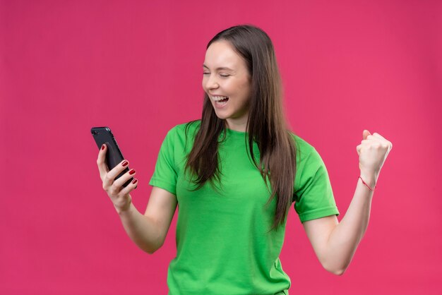 Jeune belle fille portant un t-shirt vert tenant le smartphone levant le poing fou heureux se réjouissant de son succès debout sur fond rose isolé