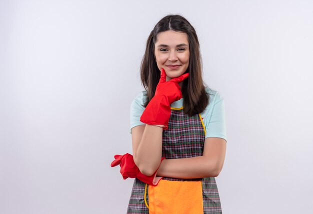 Jeune belle fille portant un tablier, une casquette et des gants en caoutchouc regardant la caméra avec la main sur le menton souriant confiant