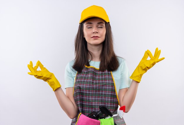 Jeune belle fille portant un tablier, une casquette et des gants en caoutchouc se détendre avec les yeux fermés faisant un geste de méditation avec les doigts