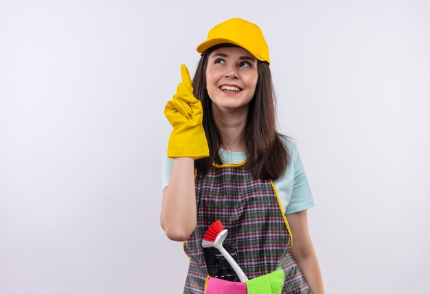 Jeune belle fille portant un tablier, une casquette et des gants en caoutchouc souriant joyeusement pointant avec le doigt