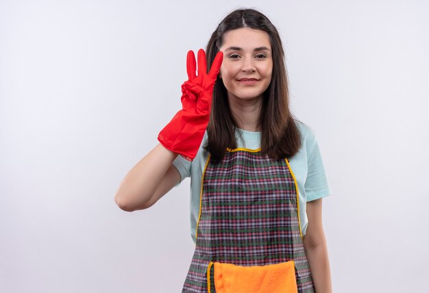 Jeune belle fille portant un tablier et des gants en caoutchouc montrant et pointant vers le haut avec les doigts numéro trois souriant
