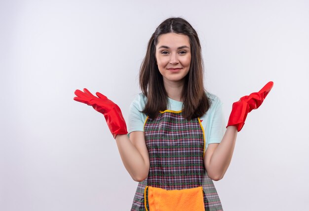 Jeune belle fille portant un tablier et des gants en caoutchouc regardant la caméra en souriant étalant les bras sur les côtés