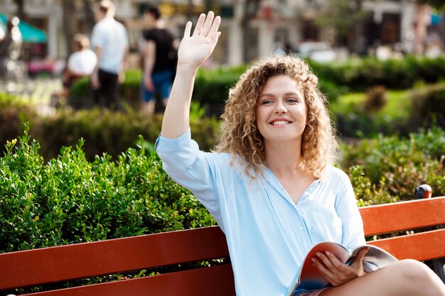 Photo gratuite jeune belle fille saluant, assis sur un banc dans le parc de la ville.