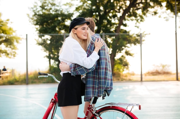 Jeune belle fille souriante aux cheveux blonds regardant joyeusement de côté tout en embrassant un garçon sur un vélo rouge sur un terrain de basket dans le parc