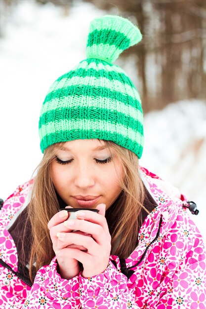 Jeune belle fille souriante dans une veste cramoisie et un chapeau vert buvant du thé chaud à partir d&#39;un thermos dans des montagnes enneigées