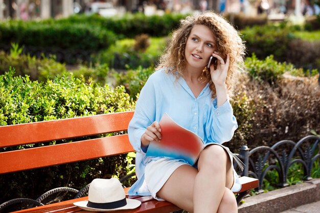 Jeune belle fille souriante, parlant au téléphone, assise sur un banc dans le parc de la ville.