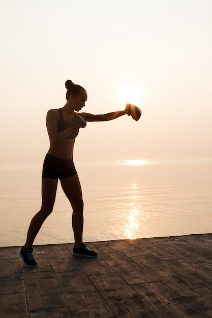Jeune belle fille sportive s'entraînant à la boxe au bord de la mer. Silhouette à l'aube.