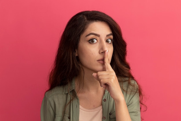 Jeune belle fille suspecte portant un t-shirt vert olive montrant un geste de silence isolé sur un mur rose