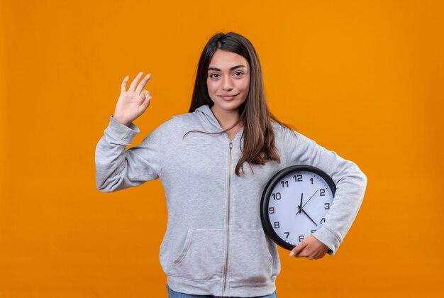 Jeune belle fille en sweat à capuche gris faisant le doigt comme le geste okey et l'autre main, elle prend l'horloge debout sur fond orange