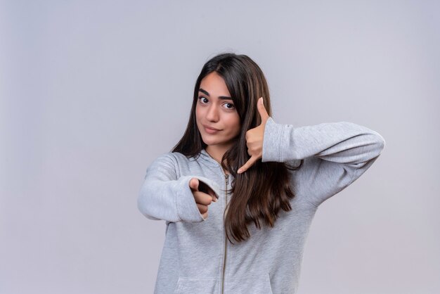 Jeune belle fille en sweat à capuche gris regardant la caméra sérieusement pointant vers la caméra faisant appelez-moi geste debout sur fond blanc