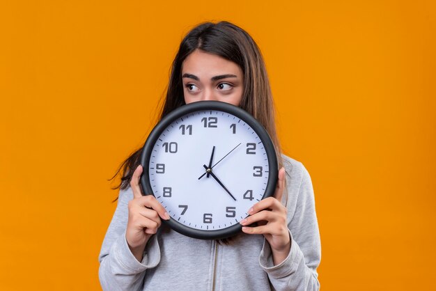 Jeune belle fille en sweat à capuche gris tenant horloge et détourner le regard avec inquiétude debout sur fond orange d