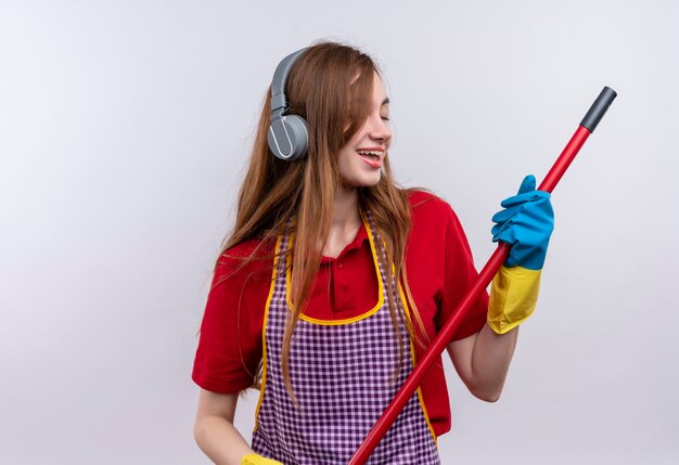 Jeune belle fille en tablier et gants en caoutchouc avec des écouteurs sur la tête tenant une vadrouille appréciant sa musique préférée