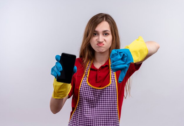 Jeune belle fille en tablier et gants en caoutchouc montrant smartphone montrant les pouces vers le bas mécontent