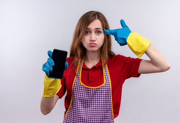 Jeune belle fille en tablier et gants en caoutchouc montrant le temple de pointage smartphone pour erreur à la confusion