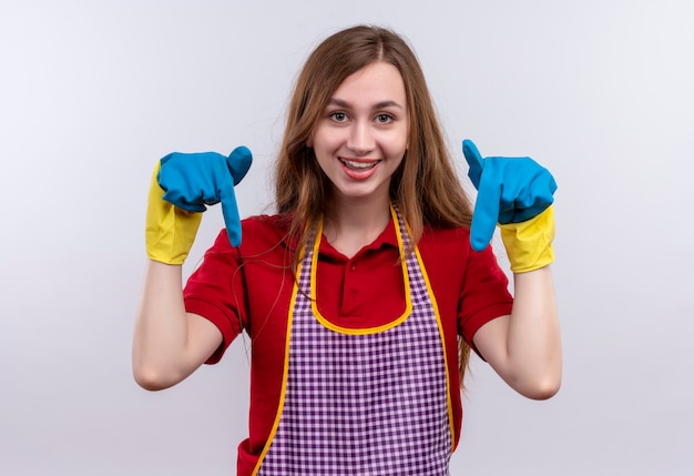 Jeune belle fille en tablier et gants en caoutchouc souriant pointant avec les doigts vers le bas