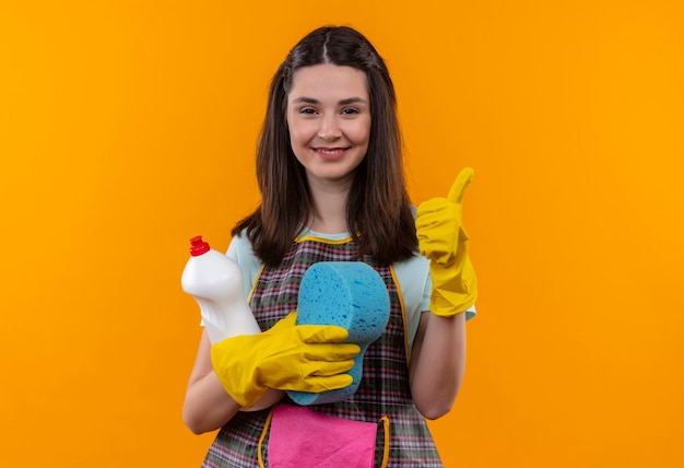 Jeune belle fille en tablier et gants en caoutchouc tenant des accessoires de nettoyage et une éponge regardant la caméra en souriant joyeusement montrant les pouces vers le haut