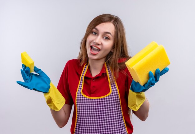 Jeune belle fille en tablier et gants en caoutchouc tenant des éponges à l'incertain et confus n'ayant pas de réponse