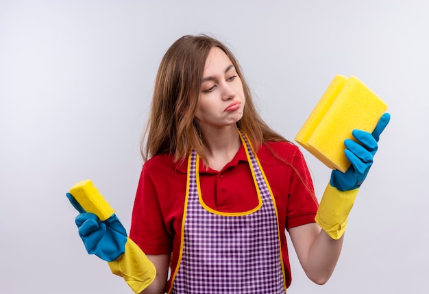 Jeune belle fille en tablier et gants en caoutchouc tenant des éponges en les regardant incertain et confus en essayant de faire un choix