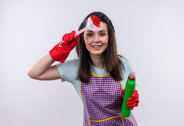 Jeune belle fille en tablier et gants en caoutchouc tenant des produits de nettoyage et une brosse à récurer à la recherche de loin avec la main sur la tête pour regarder quelque chose