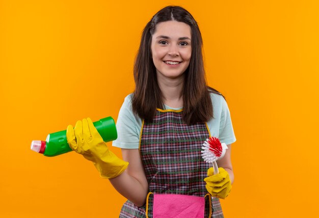 Jeune belle fille en tablier et gants en caoutchouc tenant des produits de nettoyage et une brosse à récurer souriant confiant