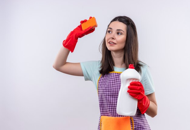 Jeune belle fille en tablier et gants en caoutchouc tenant des produits de nettoyage et une éponge souriant confiant, va nettoyer