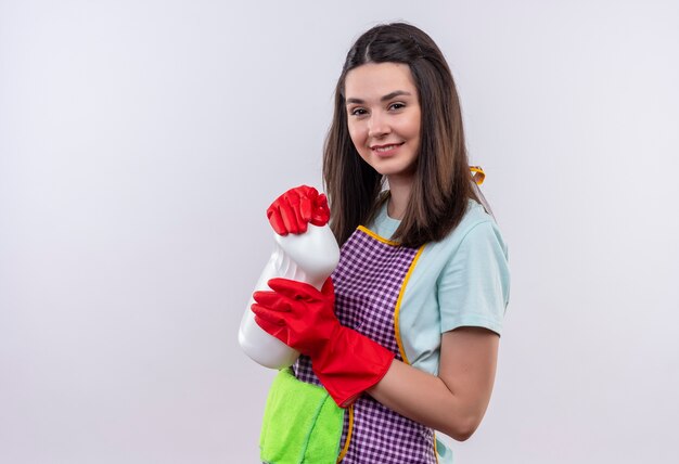 Jeune belle fille en tablier et gants en caoutchouc tenant des produits de nettoyage et un tapis regardant une caméra avec un sourire confiant