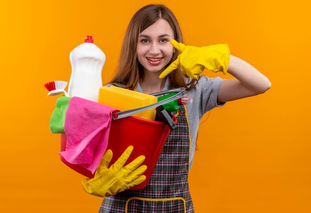 Jeune belle fille en tablier et gants en caoutchouc tenant le seau avec des outils de nettoyage pointant avec le doigt vers lui souriant joyeusement