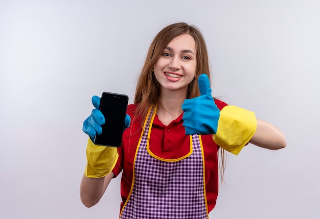 Jeune belle fille en tablier et gants en caoutchouc tenant le smartphone montrant les pouces vers le haut en souriant