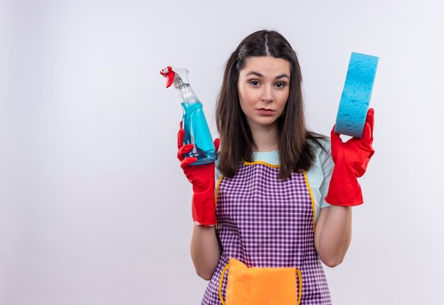 Jeune belle fille en tablier et gants en caoutchouc tenant un spray de nettoyage et une éponge regardant la caméra avec une expression sceptique