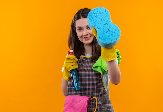 Jeune belle fille en tablier et gants en caoutchouc tenant un spray de nettoyage et une éponge regardant la caméra en souriant avec un visage heureux, prêt pour le nettoyage
