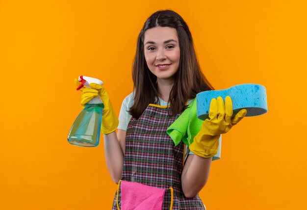 Jeune belle fille en tablier et gants en caoutchouc tenant un spray de nettoyage et une éponge regardant la caméra en souriant avec un visage heureux