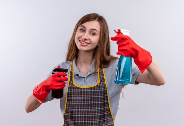 Jeune belle fille en tablier et gants en caoutchouc tenant des sprays de nettoyage souriant joyeusement