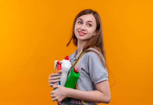 Jeune belle fille en tablier tenant des produits de nettoyage à côté souriant sympathique, prêt pour le nettoyage