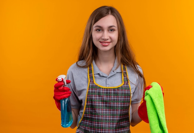 Jeune belle fille en tablier tenant spray de nettoyage et tapis regardant la caméra en souriant, prêt pour le nettoyage