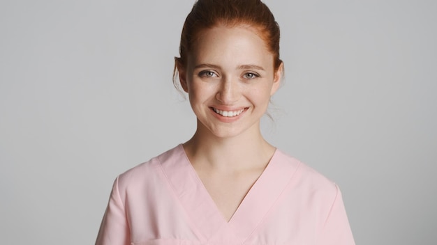 Jeune belle infirmière souriante en uniforme regardant joyeusement à huis clos sur fond blanc