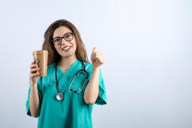 Jeune belle infirmière avec stéthoscope tenant une tasse de café et montrant un pouce vers le haut