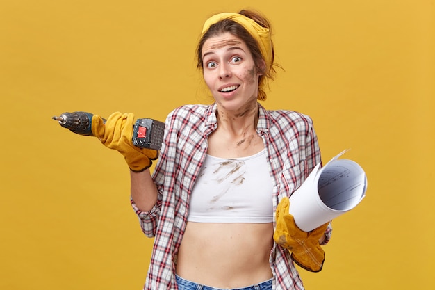 Photo gratuite jeune belle travailleuse du service de maintenance tenant la perceuse et le plan portant chemise et haut blanc ayant un regard douteux en haussant les épaules. concept de personnes, de profession et d'occupation.