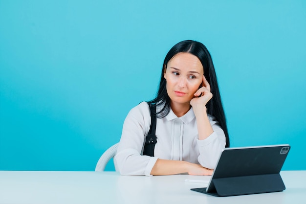 Jeune blogueuse fille pose à la caméra de la tablette en tenant la main sur la joue sur fond bleu