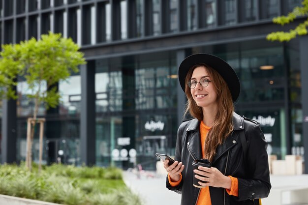 Jeune blogueuse en tenue élégante utilise un téléphone portable moderne et une connexion Internet gratuite pour la recherche de site Web