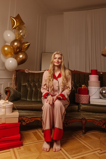 Jeune blonde caucasienne aux cheveux longs en pyjama regarde la caméra assise sur un canapé dans une chambre décorée de ballons Concepts de fête d'anniversaire