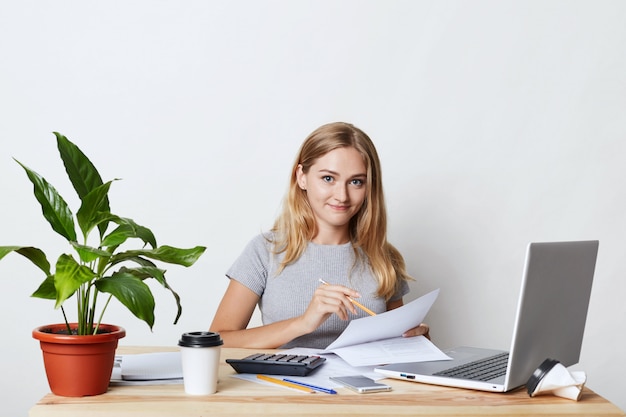 Jeune blonde femme d'affaires assise sur son lieu de travail tout en faisant un rapport d'activité, en calculant des chiffres annuels, en lisant des documents et en utilisant les technologies modernes pour son travail, en buvant du café