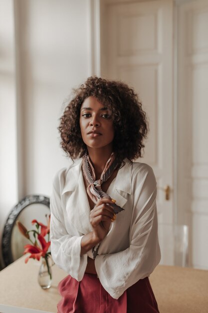 Jeune brune belle femme bouclée en pantalon bordeaux et chemisier blanc regarde devant, se penche sur une table en bois dans une chambre confortable