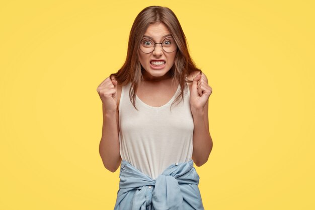 Jeune brune en colère avec des lunettes posant contre le mur jaune