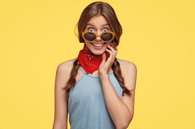 Jeune brune à la mode avec des lunettes de soleil posant contre le mur jaune