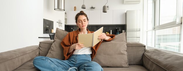 Une jeune brune souriante est assise sur le canapé du salon, tient un cahier, lit ses notes, étudie.