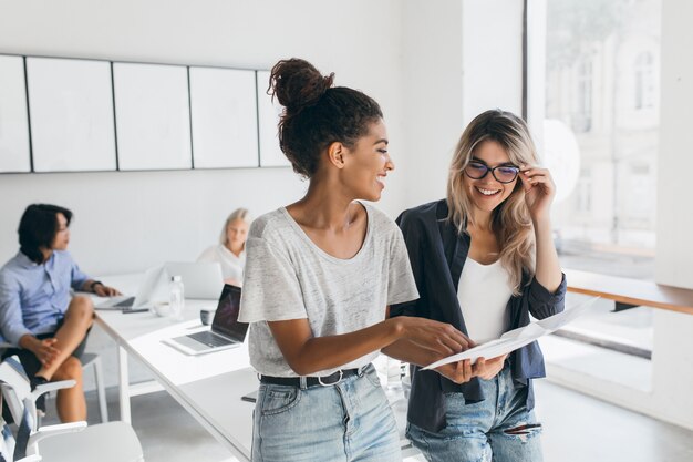 Jeune cadre féminin explique la nouvelle stratégie à l'employé blond dans des verres et souriant. Portrait intérieur d'un collectif multiculturel travaillant sur un projet au bureau et utilisant un ordinateur portable.