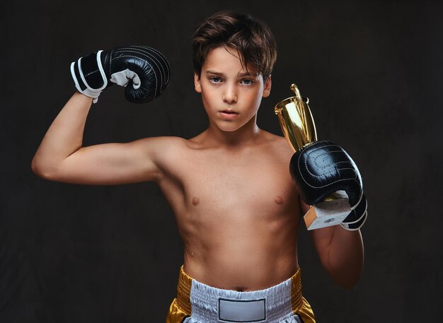 Le jeune champion de boxeur torse nu portant des gants tient une coupe du gagnant montrant les muscles. Isolé sur un fond sombre.